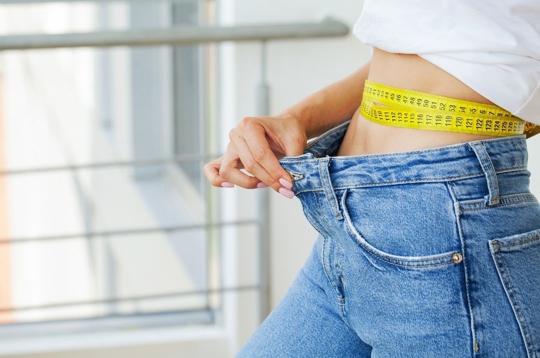 Kaloriendefizit dargestellt von schlanker Frau mit XXL Jeans und Maßband um ihren Bauch